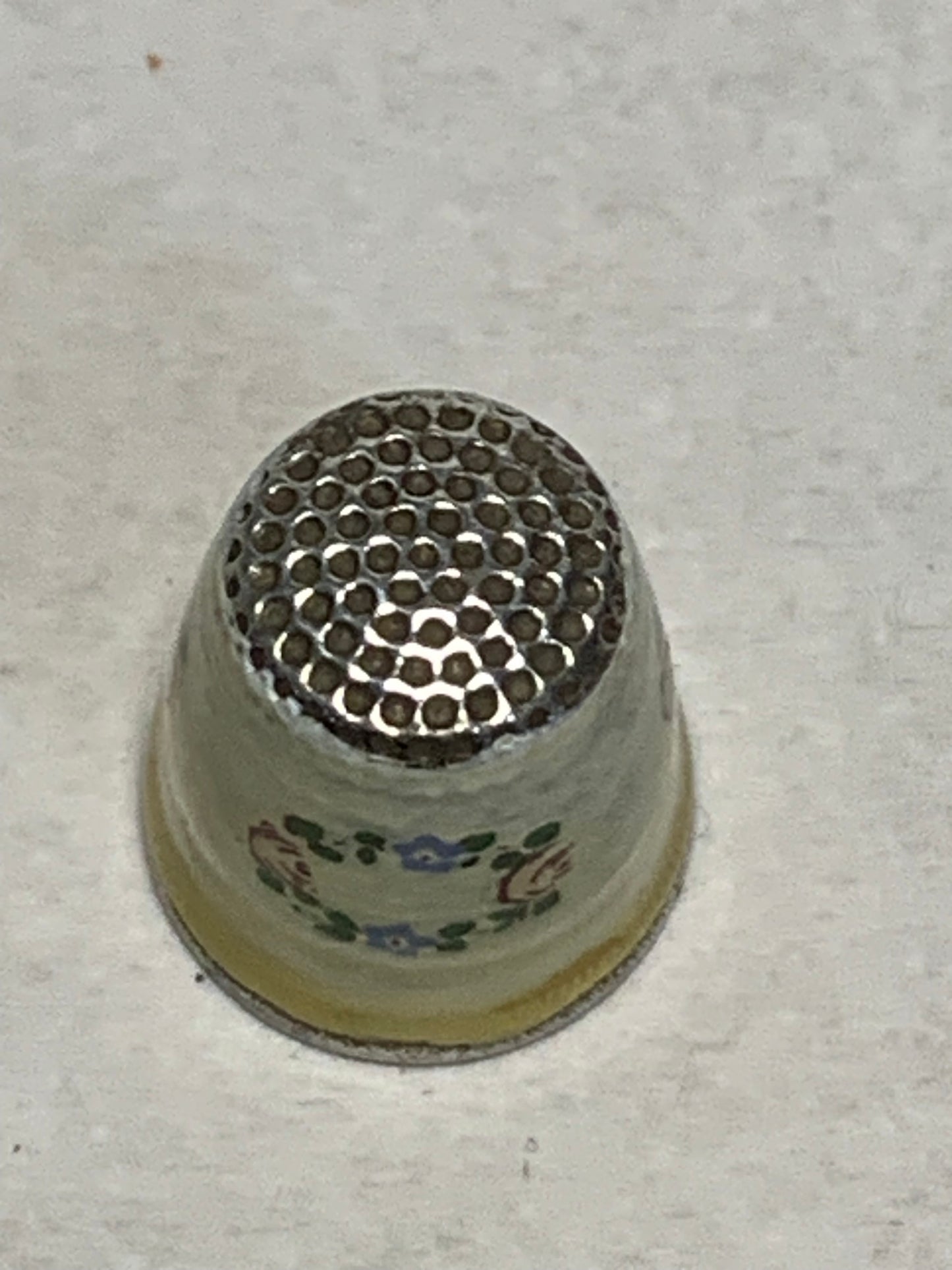 Antique metal thimble with hand-painted enamel floral decoration — a wreath of pink roses and blue forget-me-nots with green leaves encircling the body, set against a cream/ivory enameled ground