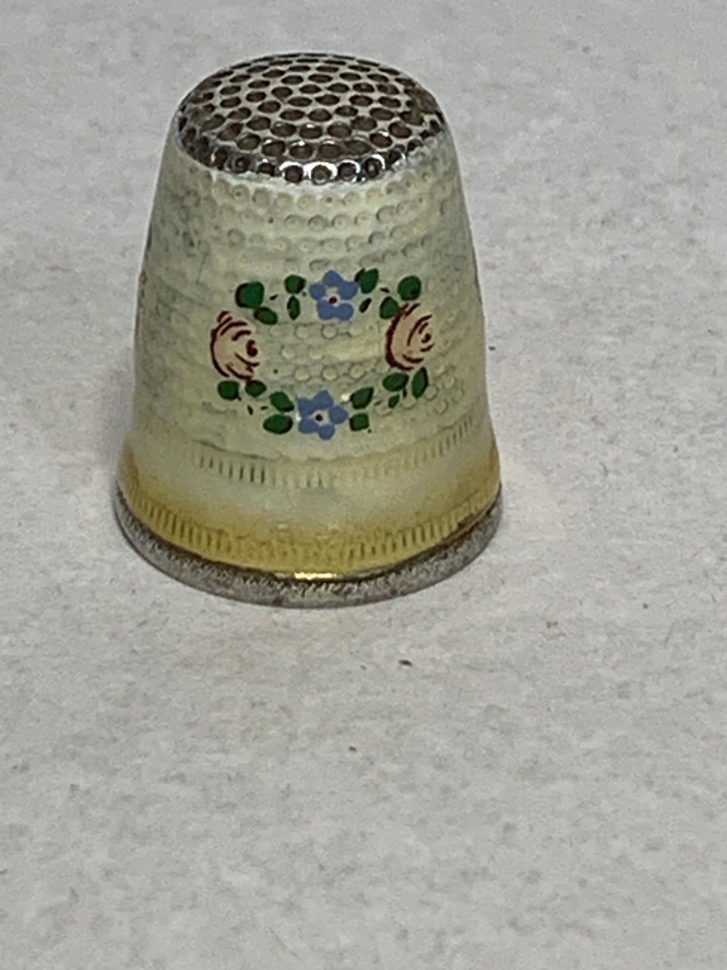 Antique metal thimble with hand-painted enamel floral decoration — a wreath of pink roses and blue forget-me-nots with green leaves encircling the body, set against a cream/ivory enameled ground