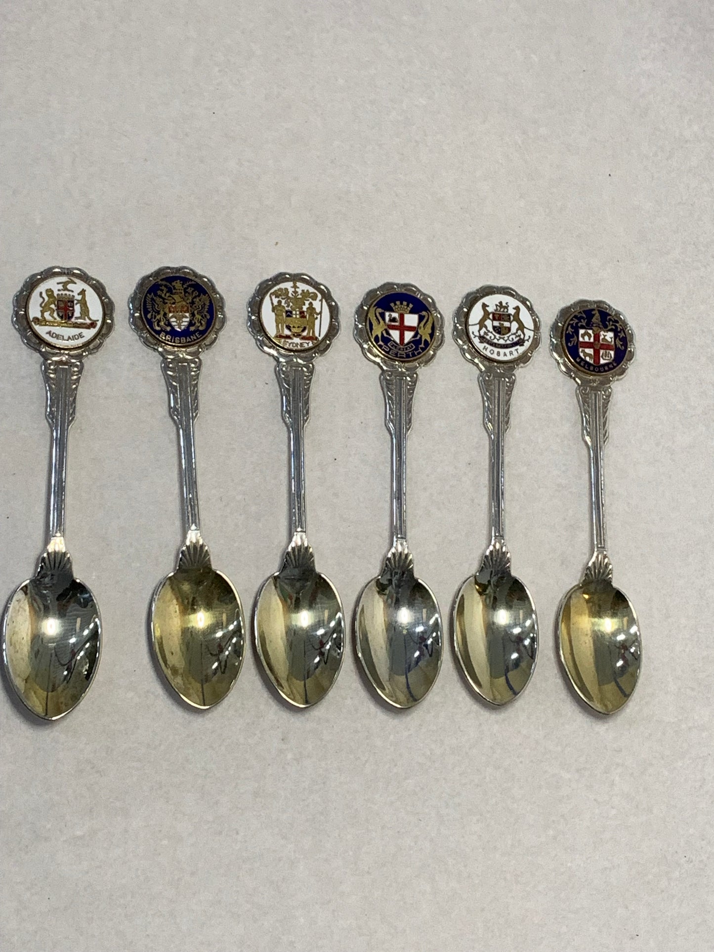 Set of six vintage silver-plated souvenir teaspoons featuring the enameled coats of arms of Australia’s state capital cities: Adelaide, Brisbane, Sydney, Perth, Hobart, and Melbourne