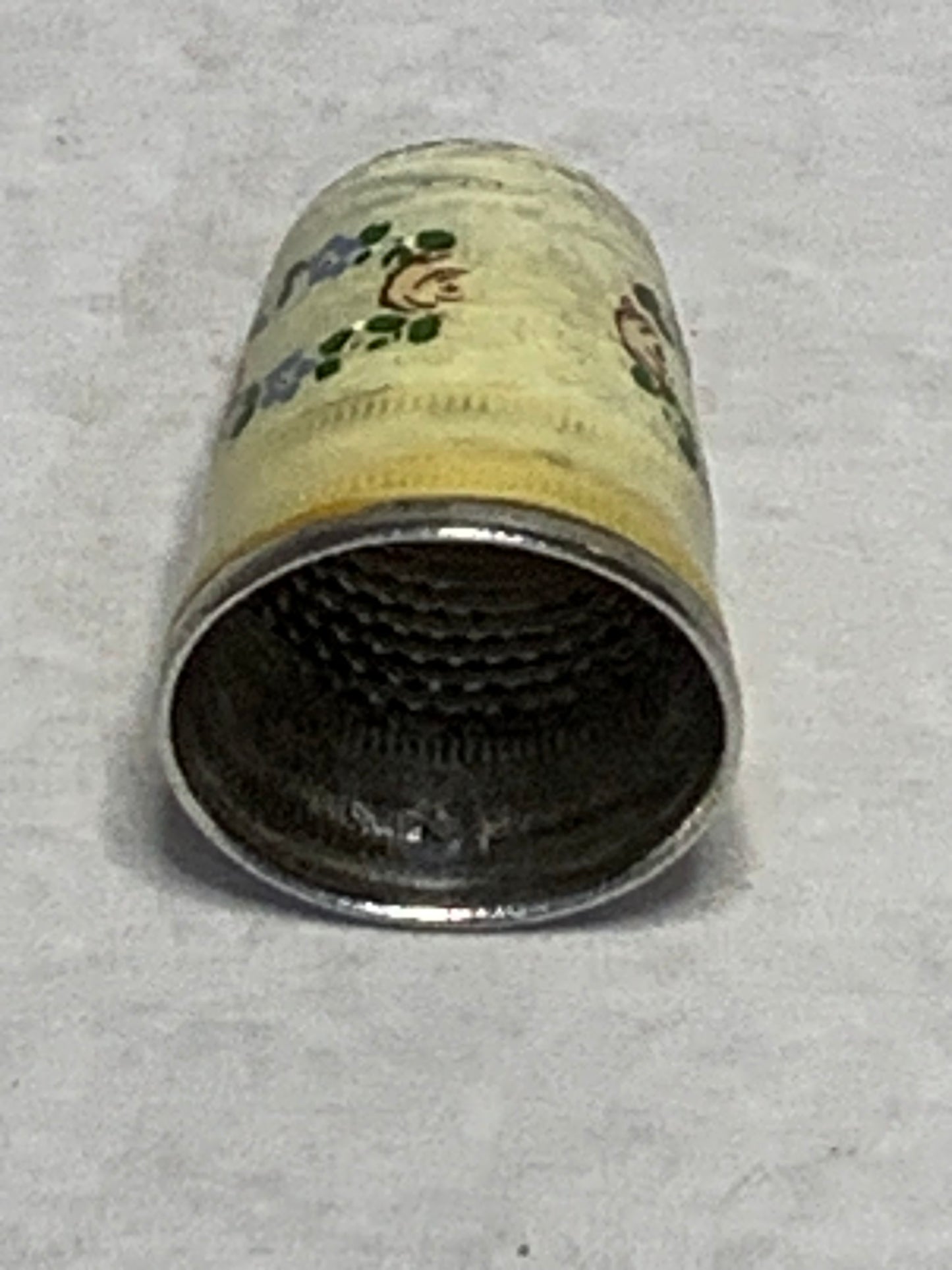 Antique metal thimble with hand-painted enamel floral decoration — a wreath of pink roses and blue forget-me-nots with green leaves encircling the body, set against a cream/ivory enameled ground